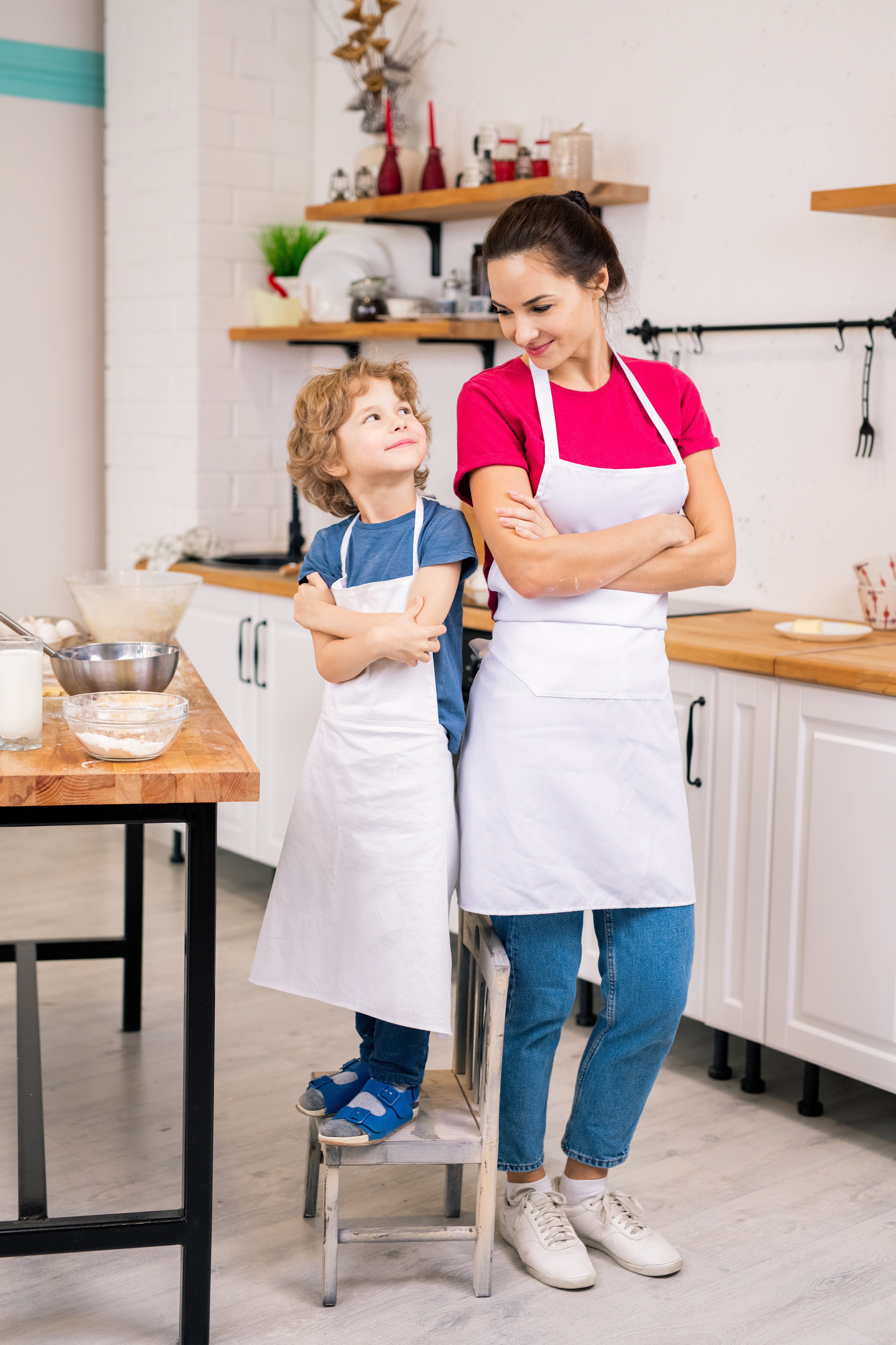 Happy adorable boy and his mother in white aprons crossing arms on chest while looking at one another in the kitchen